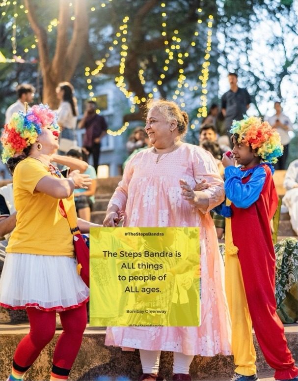 Clowns in rainbow wigs dancing with a smiling older woman, beside a sign reading The Steps Bandra is all things to people of all ages