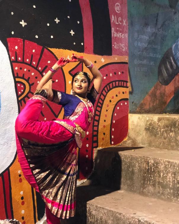 A dancer in a pink sari posing against a bold red and gold mural